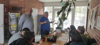 two persons standing in a room and talking, five persons sitting around a table and listening
