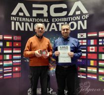 Two teachers with an award certificate in front of a ARCA International Exhibition of Innovation billboard