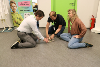 Three people sitting on the floor and working on a small wooden model 